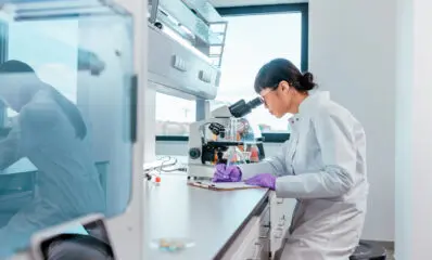 A female scientist is sitting in her lab and looking trough the microscope, she is writing down notes about her discoveries after looking at samples